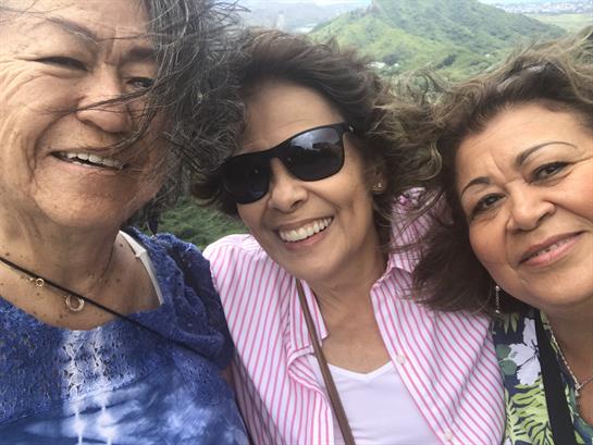 Three friends pose for a cheerful selfie with green mountains and a bright sky behind them.