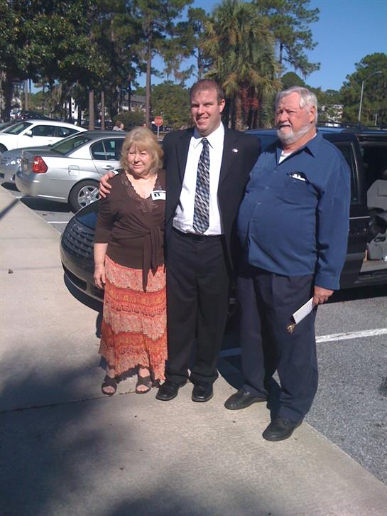 Three individuals are standing together in a parking lot near parked cars, smiling cheerfully.