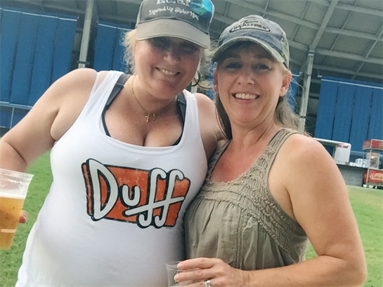 Two friends smile and pose together, holding drinks under a large venue structure.