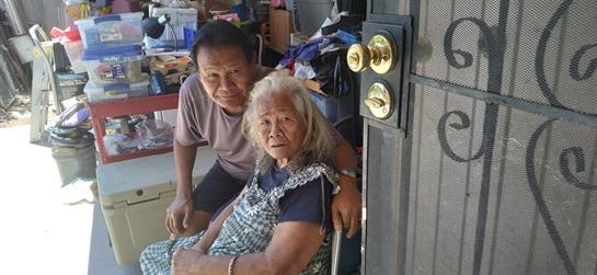 A man and an elderly woman smile while sitting together by a home entrance in bright daylight.