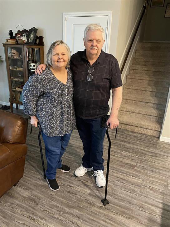 An elderly man and woman pose together in a welcoming living room while using mobility aids.
