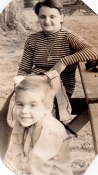 A smiling child sits in the grass next to a woman at a picnic table in nature.