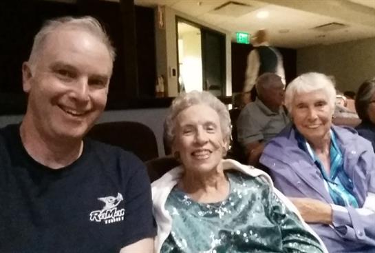 Two seniors smile cheerfully alongside a man as they enjoy an evening gathering together.
