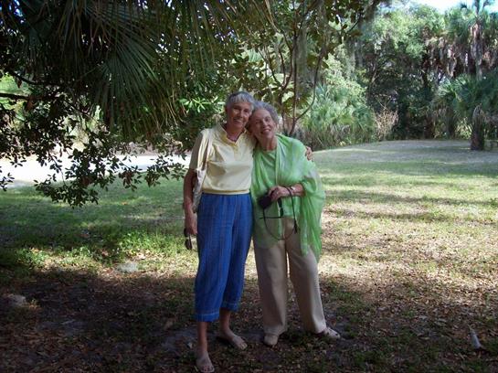 Two elderly women stand close together in a park, enjoying a sunny day surrounded by greenery.