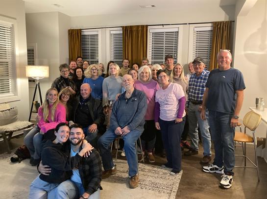 A large group of friends and family smiles while posing together in a warm living room.
