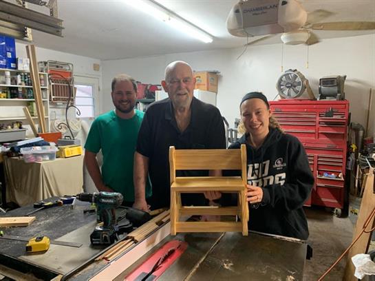 Three people proudly display a wooden step stool they created together in a workshop.