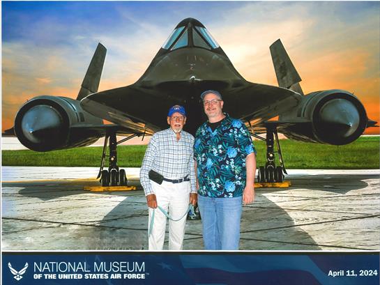 Two men stand together, smiling in front of a dark military aircraft in a museum.