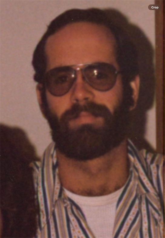 A man wearing sunglasses and a striped shirt stands indoors displaying a serious demeanor.