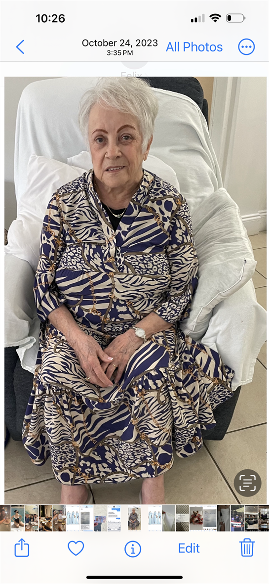 An elderly woman sits in a comfortable chair, wearing a colorful patterned dress, looking content.