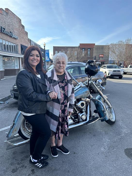 A young woman stands beside an elderly woman next to a classic motorcycle in town.