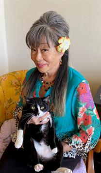 Elderly woman smiles while holding a black and white cat in her lap, wearing vibrant clothing.