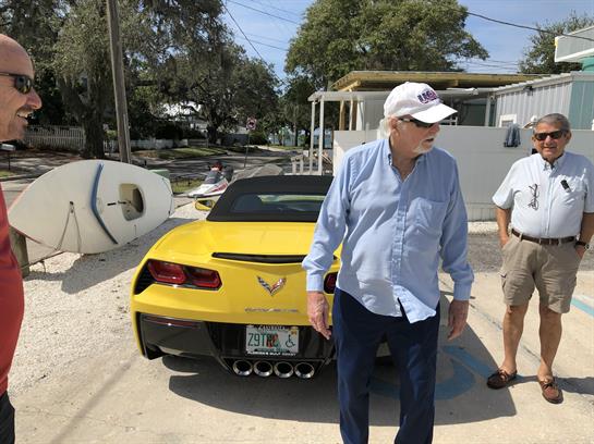 Three men talk casually next to a bright yellow sports car in a coastal area.