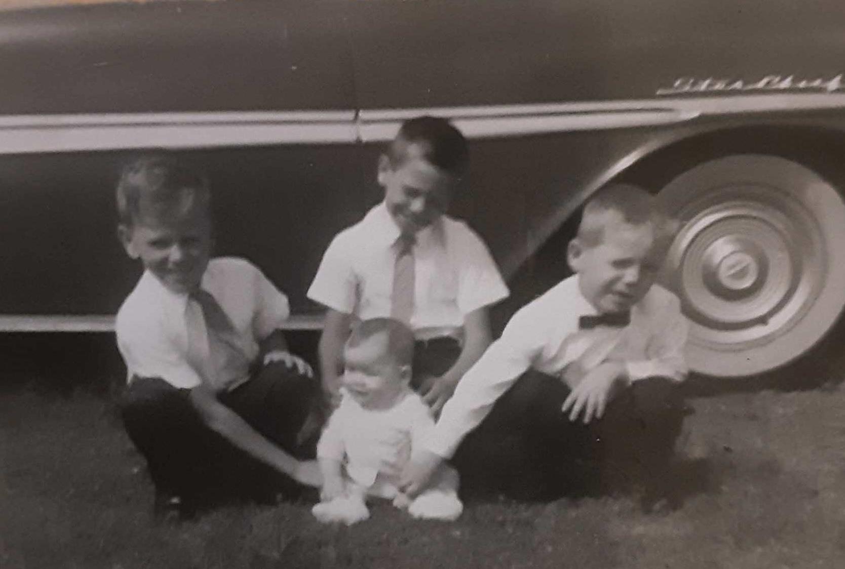 Four young boys sit on the grass, enjoying time together beside a classic automobile.