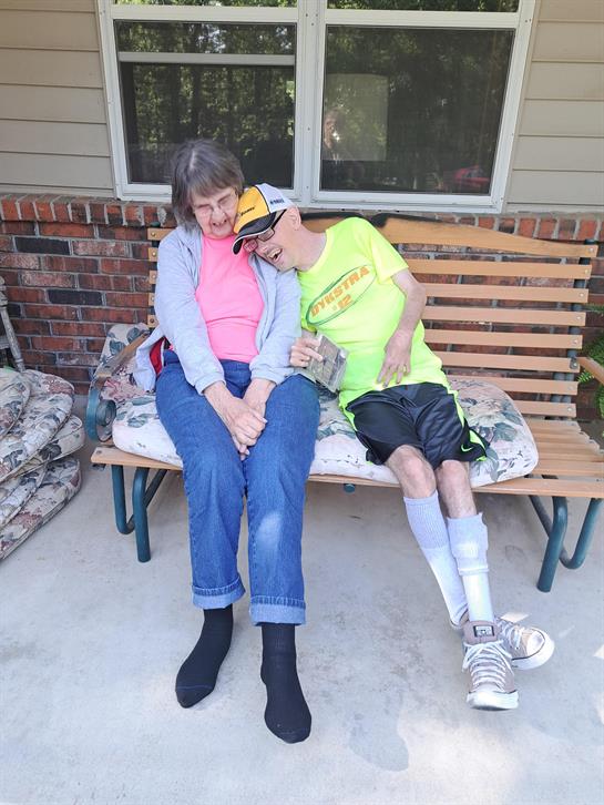 Two individuals share a moment on a bench outside, relaxed and smiling together in sunlight.