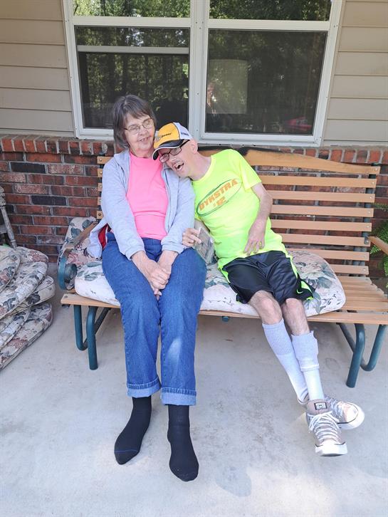 Two individuals enjoy each other's company on a bench outside a house in the sun.