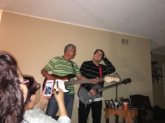 Two men perform music together with guitars while family members watch and enjoy the fun.