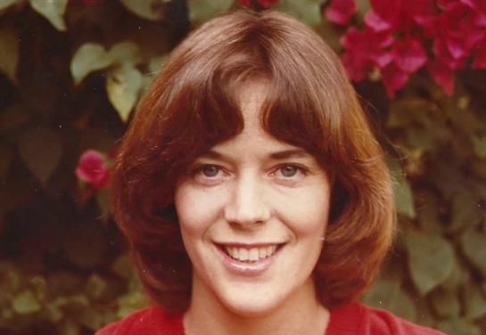 A woman with shoulder-length hair smiles warmly amidst colorful flowers, radiating joy.
