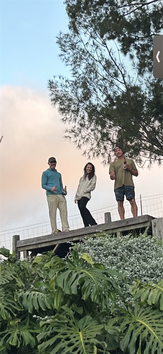 Friends pose happily on a wooden deck with lush plants and a clear sky above.