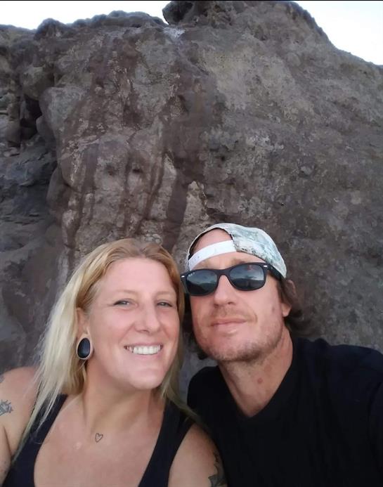 A couple poses happily while capturing a moment at a beach by rocky formations.