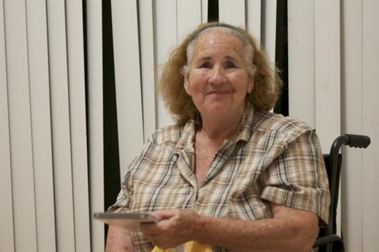 A smiling elderly woman in a wheelchair holds a plate of food while enjoying a gathering.