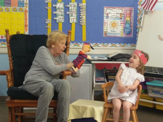 A senior woman engages a young girl with a puppet during a playful storytelling session.