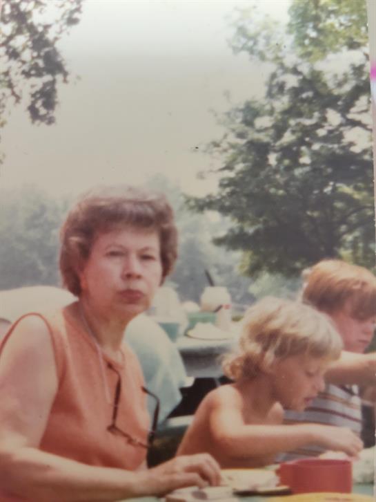 A woman observes children at a summer picnic while enjoying food in a serene outdoor setting.