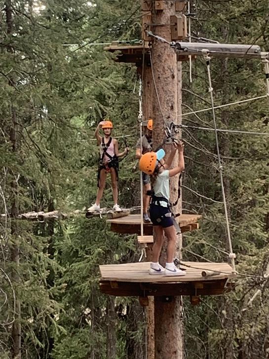 Kids navigate obstacles in a tree-top course while wearing climbing gear and helmets.