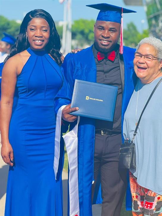 A proud graduate poses with family members, smiling widely at a sunny outdoor graduation event.