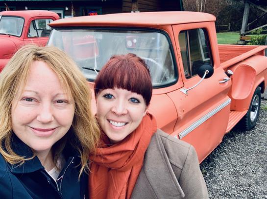 Two women smile for a selfie beside a classic red pickup truck in a rustic outdoor setting.