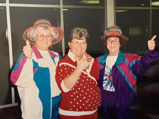 Three women in vibrant outfits share smiles and thumbs-up gestures at a cheerful gathering.