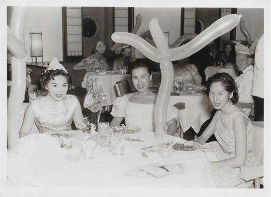 Three women in elegant attire smile brightly while seated at a table adorned with balloons.