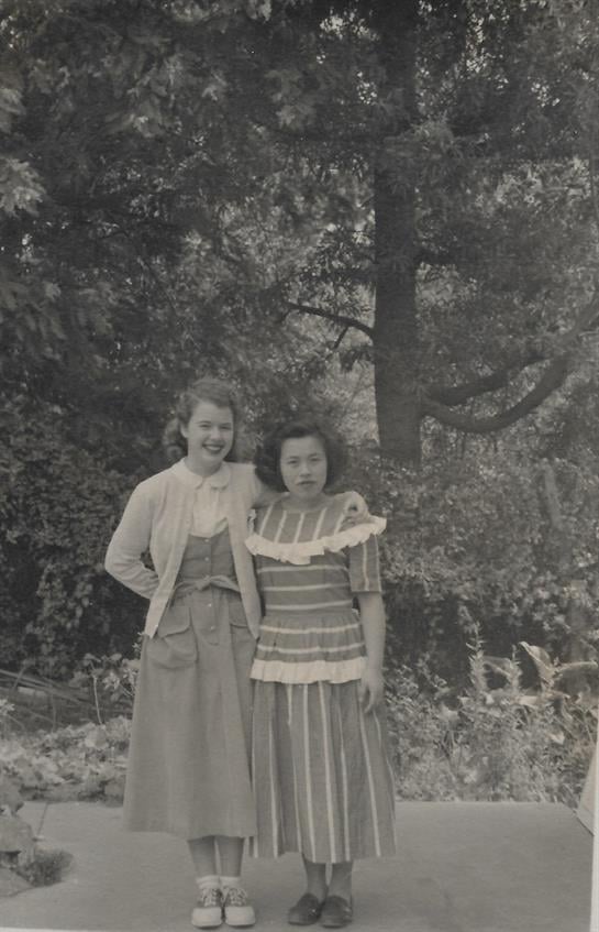 Two friends stand in a lush garden, smiling and enjoying each other's company on a sunny day.