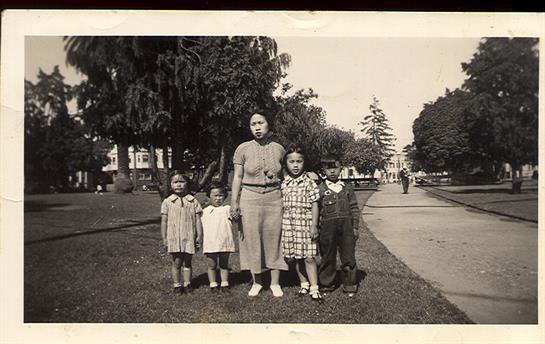 Five children stand in a park with an adult, surrounded by green trees and pathways.