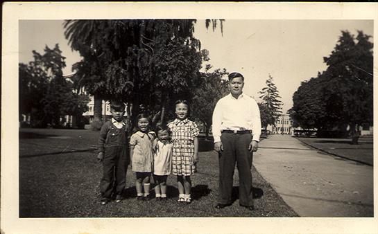 Group of four children and an adult man pose for a picture in a lush park filled with trees.