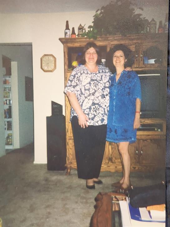 Two friends stand side by side in a cozy living room, smiling warmly at the camera.