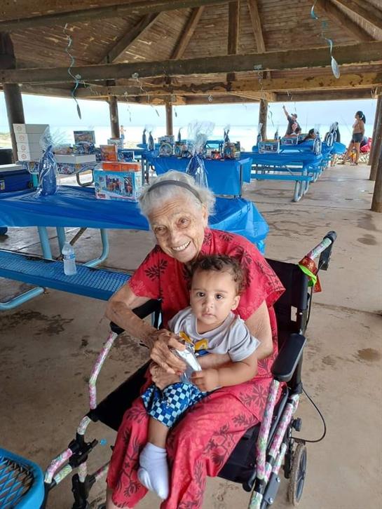 An elderly woman in a wheelchair joyfully holds a toddler during a family gathering.