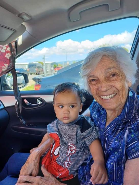 An elderly woman smiles while holding a toddler in a car. The child looks curiously outside.