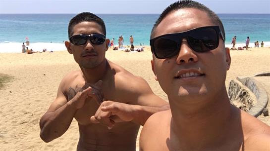 Friends smile and pose for a selfie on a sunny beach with clear blue water in the background.