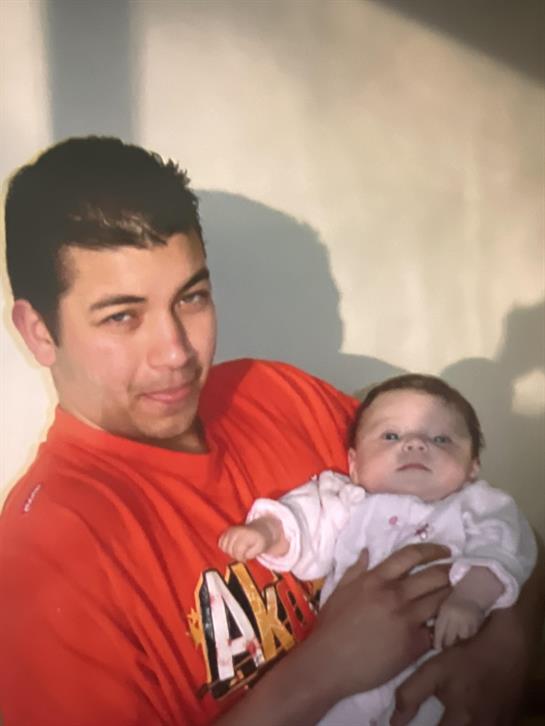A young man with dark hair smiles while holding a baby in a cozy indoor space.
