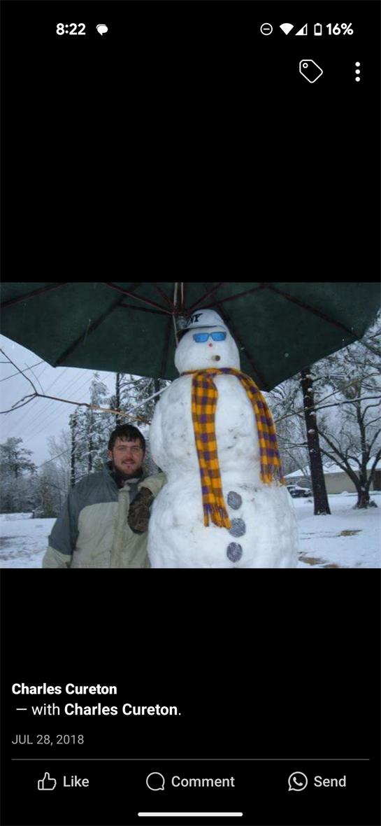 A man stands proudly next to a large snowman with a scarf and umbrella in a winter scene.