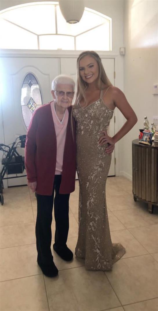 A young woman in a glamorous gown stands with an elderly woman in a cozy home.