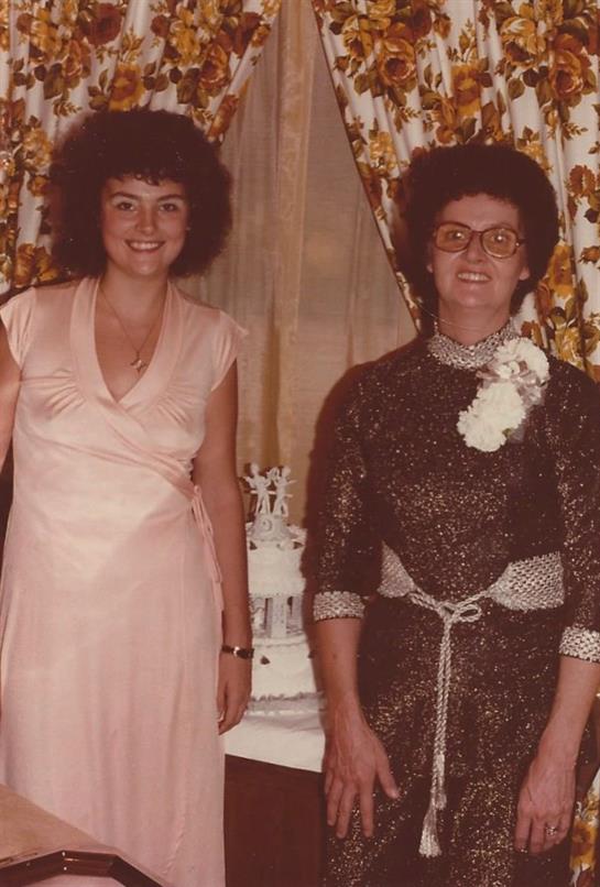 Two women dressed in formal attire stand together smiling in a warmly decorated room.