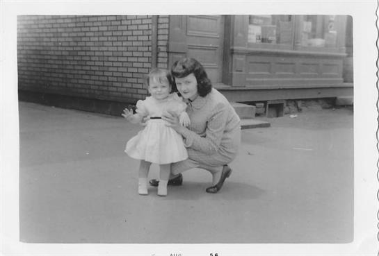A mother kneels beside her young daughter, both smiling, in a quaint outdoor setting.