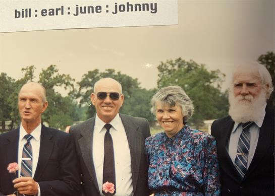 Four adults stand together in formal wear, holding small flowers, smiling during a summer event.