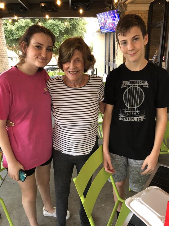 Teenagers stand together with an elderly woman, smiling in an outdoor dining area.