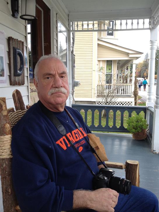 An older man sits comfortably on a porch chair, holding a camera, enjoying the sunny afternoon.
