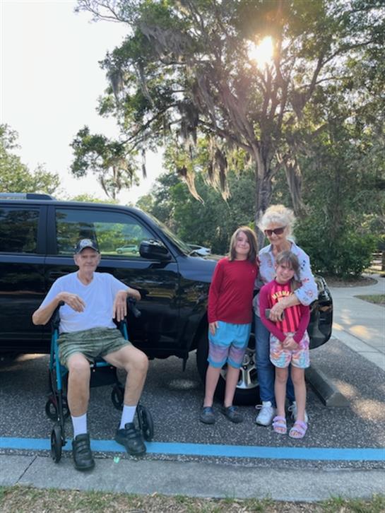 A joyful group of family members enjoys time together near a parked vehicle on a sunny afternoon.