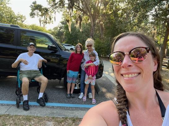 A cheerful family stands near a parked vehicle, smiling on a sunny day in a park.