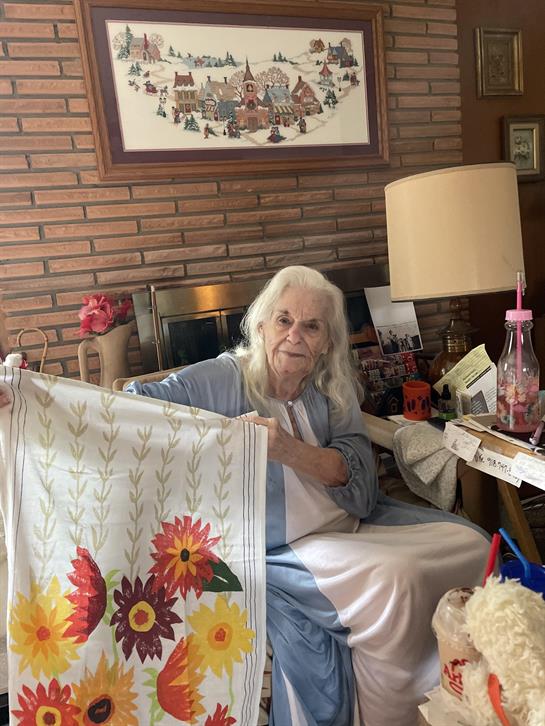 An elderly woman proudly displays her vibrant floral tablecloth in a warm, inviting living space.