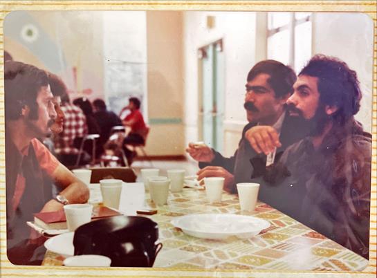 Three men interact animatedly at a table filled with cups while others sit in the background.